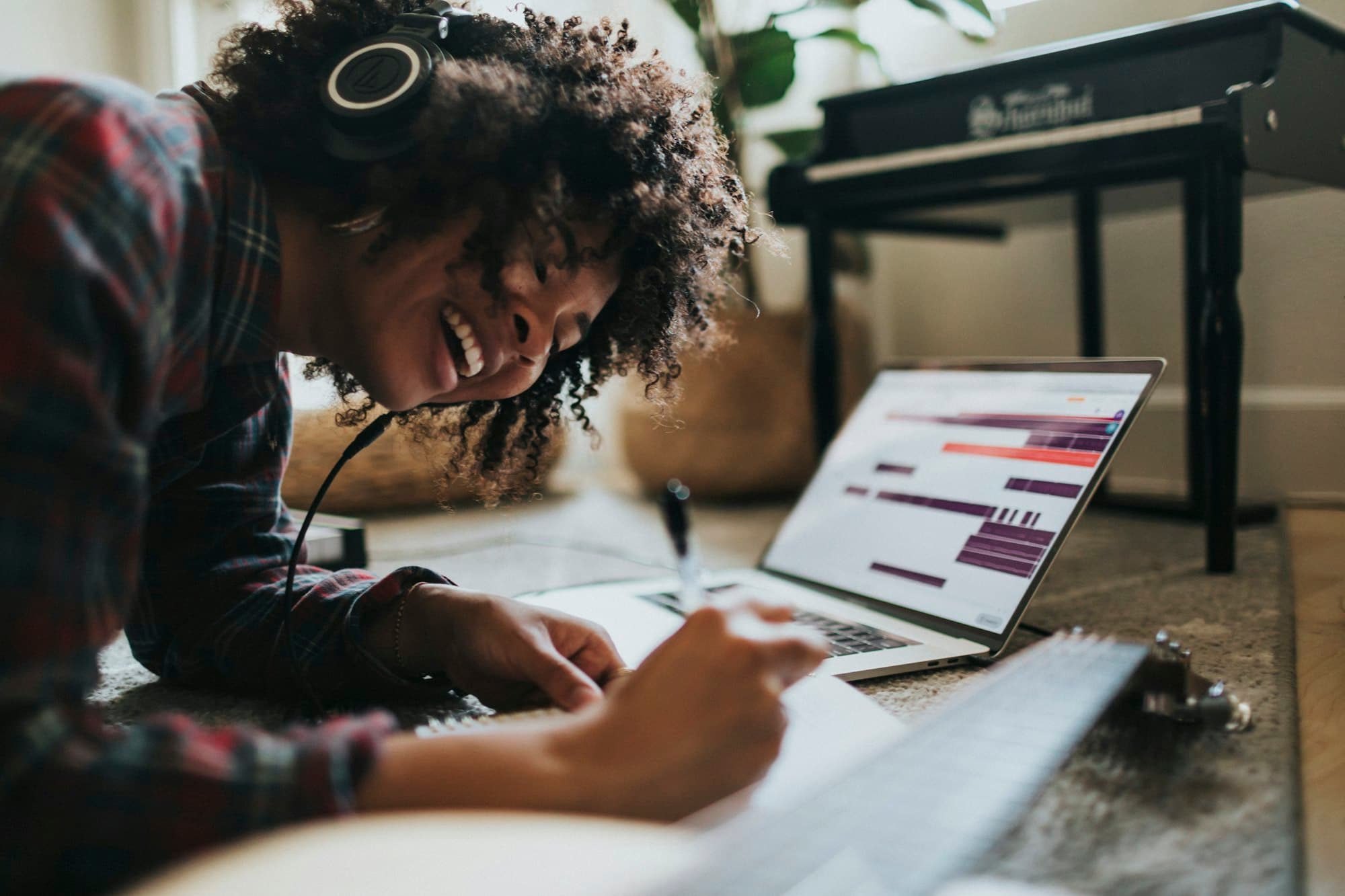 A songwriter with headphones on, smiling as she writes notes on paper beside a laptop